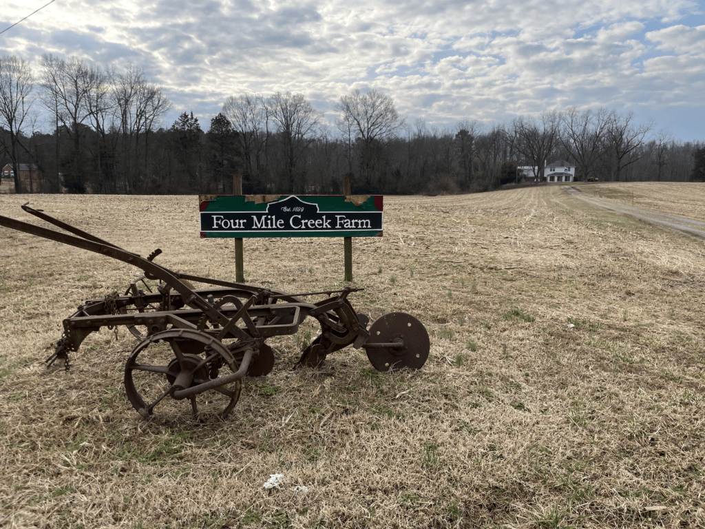 View of fields and farmhouse at Four Mile Creek Farm from New Market Road with antique plow and farm sign in foreground.